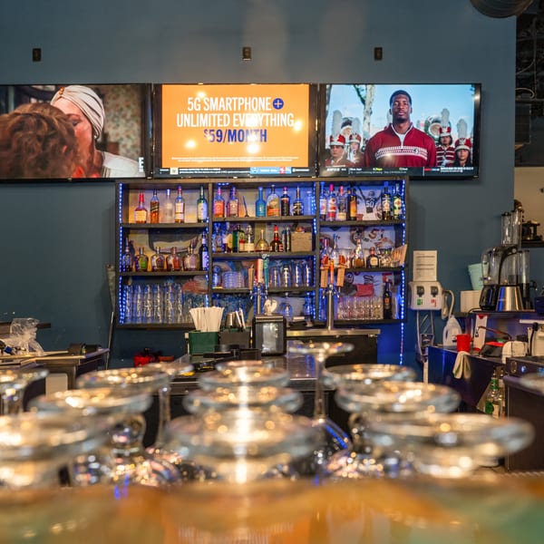 A bar area with three large TVs displaying various content, liquor bottles on shelves, and empty glasses.