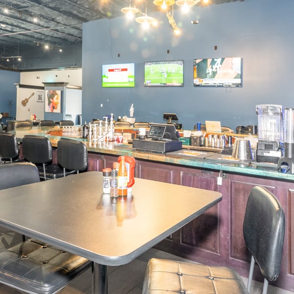 Interior of a restaurant bar with stools, a table with condiments, and three TVs showing sports.