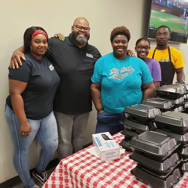 Five smiling people stand behind a table laden with stacked to-go containers and a box of gloves.