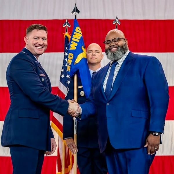 Two men, one in military uniform and one in a blue suit, shake hands in front of flags.