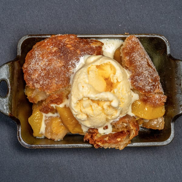 Warm peach cobbler with a scoop of melting vanilla ice cream in a dark cast iron dish.