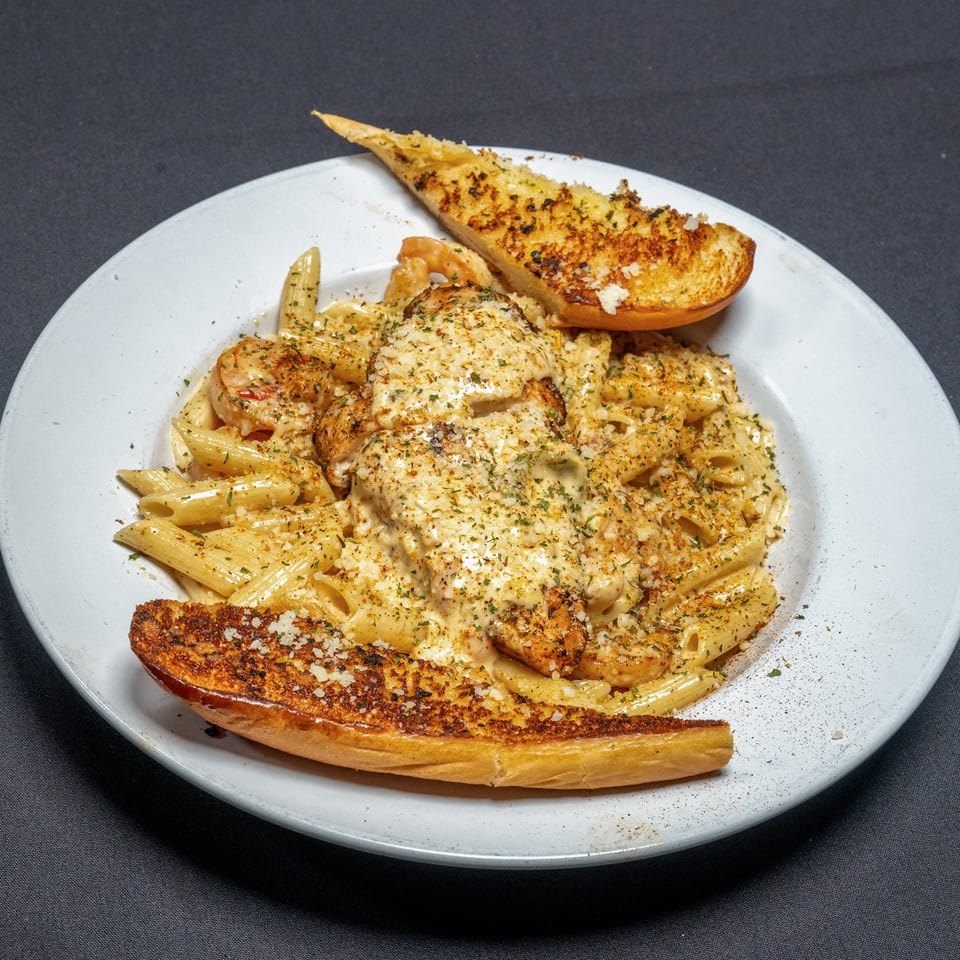 Creamy penne pasta with chicken, shrimp, and two slices of garlic bread on a white plate.
