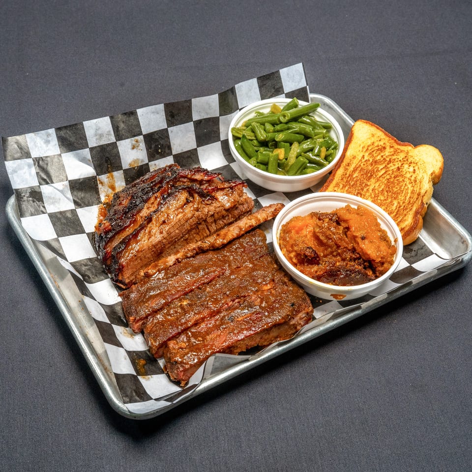 Barbecue platter with sliced brisket, ribs, green beans, sweet potatoes, and Texas toast on a tray.