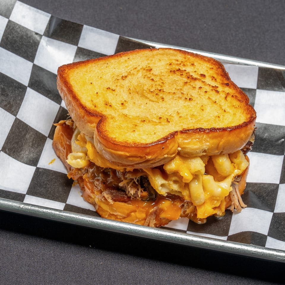 Grilled cheese sandwich filled with pulled pork, macaroni and cheese, and jalapeños on checkered paper.