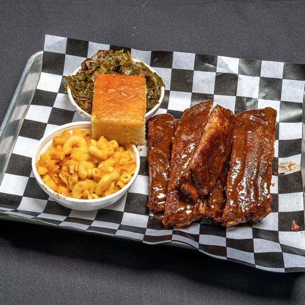 BBQ ribs, mac and cheese, collard greens, and cornbread on a metal tray with checkered paper.