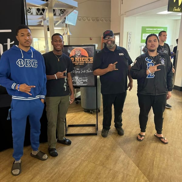 Four men pose with a "Big Nick's Soulfood-Wings" sign in a brightly lit indoor space.