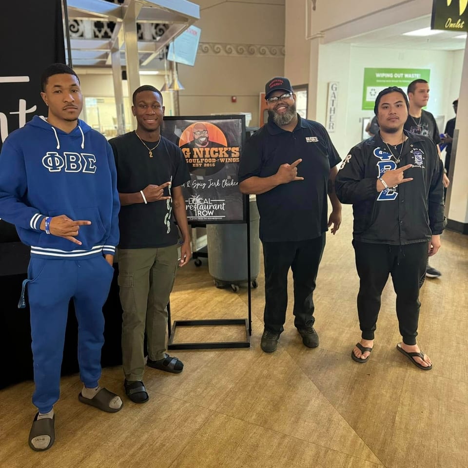Four men pose with a "Big Nick's Soulfood-Wings" sign in a brightly lit indoor space.