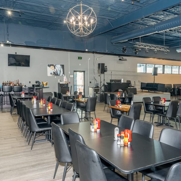Spacious restaurant interior with black tables and chairs, a bar, and a stage area.