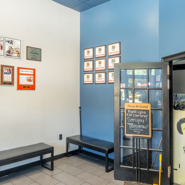 Restaurant entrance with awards on the wall, benches, and a "Please Be Seated" sign on the door.